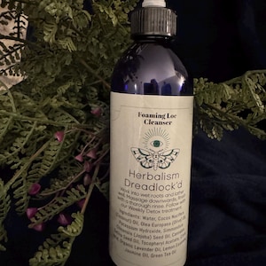 May include: A dark blue bottle of Herbalism Dreadlock'd Foaming Loc Cleanser with a white label. The label features text and a moth illustration. The bottle has a white applicator tip and is surrounded by green foliage.