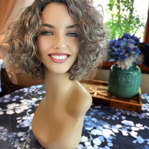May include: A mannequin head with a short, curly wig in shades of brown and blonde. The mannequin has a neutral skin tone and is positioned on a table with a blue and white floral pattern. The background includes a window and a vase of blue flowers.