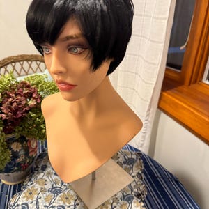 May include: A black short bob wig on a mannequin head. The wig has a straight, blunt cut and is styled with a side part. The mannequin is wearing a light brown skin tone.