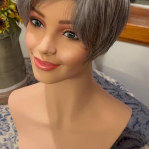 May include: A short, silver-grey wig with a side part and a choppy, layered style. The wig is styled on a mannequin head.