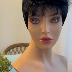 May include: A mannequin head with short, black, choppy hair. The mannequin has a neutral expression with green eyes and pink lips.