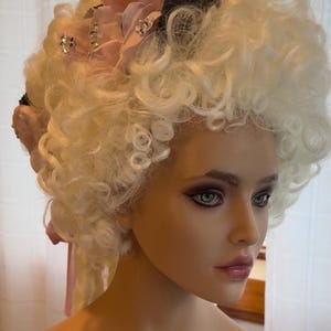 May include: A white curly wig with a pink and black flower embellishment. The wig is styled in a large, voluminous shape with curls throughout.