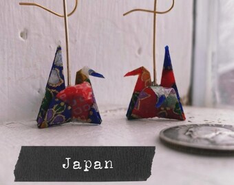 May include: A pair of origami crane earrings with gold hooks. The cranes are made of colorful patterned paper and are about 1 inch tall. The word "Japan" is written on a black background below the earrings.