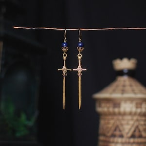 May include: A pair of gold-colored sword earrings with dark blue beads. The earrings are suspended from a thin wire against a dark background. The swords have intricate details and are a unique accessory.