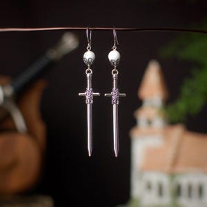 May include: A pair of silver-colored sword earrings with white beaded accents. The earrings have a detailed design on the sword's hilt and a simple hook closure. The background is blurred, with hints of greenery and a building.