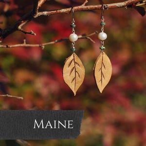 May include: A pair of handcrafted earrings featuring leaf-shaped pendants. Each earring has a light brown leather leaf, a white pearl, and green beads. The word "MAINE" is displayed on a dark gray banner at the bottom.