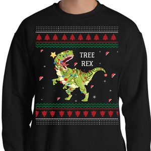 May include: Black Christmas jumper featuring a green dinosaur wearing Christmas lights and a star on its head. The text "TREE REX" is printed on the jumper.