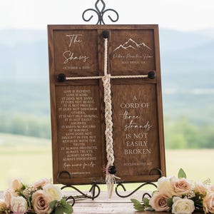 May include: Wooden cross-shaped wedding sign with the text "The Shaws" and "October 4, 2025". The sign includes a quote about love and a quote that says "A cord of three strands is not easily broken".