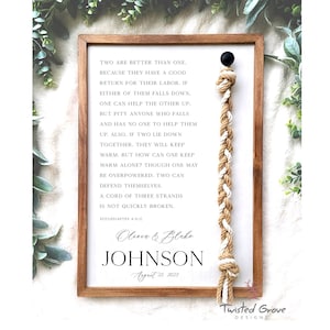 May include: Wooden framed sign with a white background and black text. The text includes a quote from Ecclesiastes and the names Olivia & Blake Johnson, with the date August 21, 2023. A decorative rope hangs from the top.