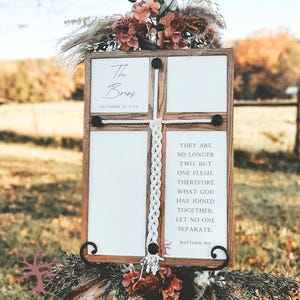 May include: A wooden wedding sign with a cross-shaped design. The sign features four white panels, with text that reads "The Brins, December 26, 2024" and a Bible verse. The sign is decorated with floral arrangements and greenery.