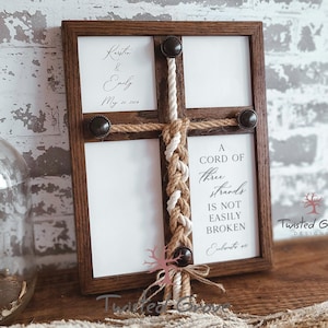 May include: A wooden frame with a white background and a braided rope in the center. The rope is a trinity braid, symbolizing three strands that are not easily broken. The frame also includes a personalized inscription with the names "Kerstin & Emily" and the date "May 20, 2024". The text "A cord of three strands is not easily broken" is also included in the frame.