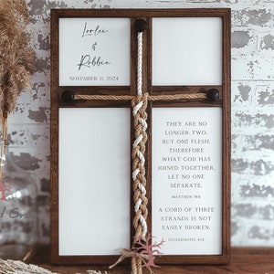 May include: A wooden sign with a white background and a brown rope braid in the center. The sign has the text "Trinity Braid" at the top, "Lorlei & Robbie" in the center, and "November 11, 2024" at the bottom. The sign also has the text "They are no longer two, but one flesh. Therefore what God has joined together, let no one separate. Matthew 19:6" and "A cord of three strands is not easily broken. Ecclesiastes 4:12" on the right side.