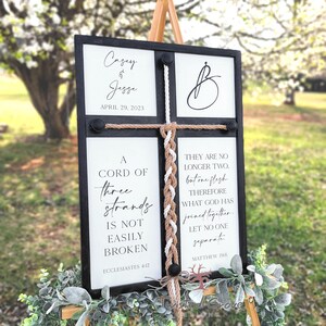 May include: A black framed sign with white text and a braided rope. The sign reads "A cord of three strands is not easily broken, Ecclesiastes 4:12" and "They are no longer two, but one flesh. Therefore what God has joined together, let no one separate. Matthew 19:6". The sign also has the names Casey and Jesse and the date April 29, 2023.