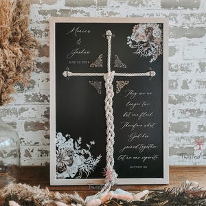 May include: A framed chalkboard sign with a rope cross, floral accents, and the text "Monica & Joshua, June 22, 2024." The sign also features a Bible verse. The sign is displayed on a wooden surface with decorative dried plants.