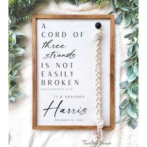 May include: A white wooden sign with the text "A cord of three strands is not easily broken, Ecclesiastes 4:12" and "TJ & Shannon Harris, November 30, 2022". A white rope with a black bead is attached to the sign.