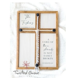 May include: A wooden sign with a white background and a brown frame. The sign has a braided rope with black beads at the ends. The sign says "A cord of three strands is not easily broken, Ecclesiastes 4:12". The sign also has a tree silhouette and the text "The Fishers, November 5, 2021" and "Twisted Grove Designs".