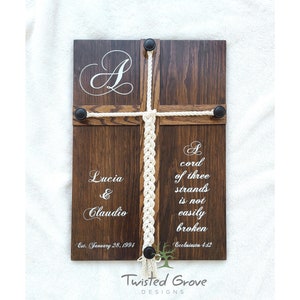 May include: A wooden plaque with a white rope braid in the center. The plaque has the text "A cord of three strands is not easily broken" and "Ecclesiastes 4:12" on it. The plaque also has the text "Lucia & Claudio" and "Est. January 28, 1994" on it. The plaque has a white initial "A" at the top and the text "Twisted Grove Designs" at the bottom.