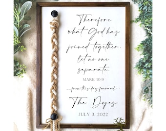 May include: A wooden sign with a white background and black text. The text reads "Therefore what God has joined together, let no one separate. Mark 10:9 from this day forward The Dyes July 3, 2022". A brown and white rope is attached to the sign with black beads at the top and bottom. The sign is framed in a dark wood frame. The text "Twisted Grove Designs" is printed below the sign.
