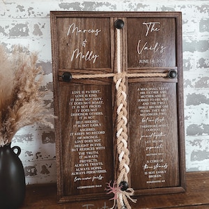 May include: A wooden framed wall art piece with a cross design. The cross is made of rope and braided twine. The artwork includes names, a date, and Bible verses in white lettering. A black vase with dried flowers is on the left.