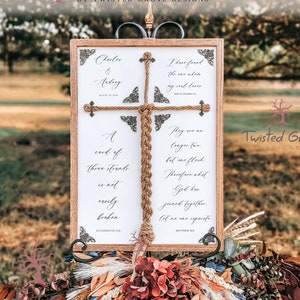 May include: A wooden sign with a rope braid in the shape of a cross. The sign has the text "Charles & Avling" and wedding date "August 11, 2014". The sign also includes two bible verses: "A cord of three strands is not easily broken" (Ecclesiastes 4:12) and "They are no longer two, but one flesh. Therefore what God has joined together, let no one separate." (Matthew 19:6).