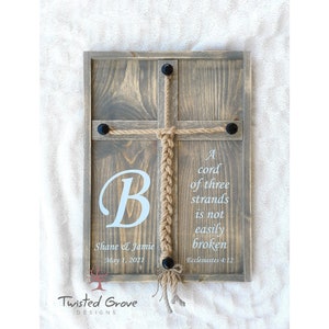 May include: A wooden sign with a braided rope in the shape of a cross. The sign has the text "A cord of three strands is not easily broken" and "Ecclesiastes 4:12". The sign also has the text "B" and "Shane & Jamie May 1, 2021".