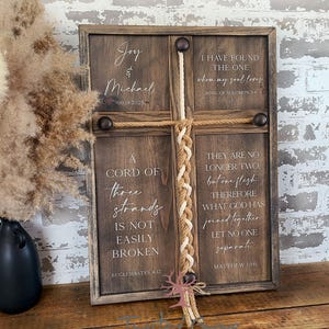 May include: A wooden cross-shaped sign with a dark brown finish. The sign features the names "Joy & Michael" and the date "09.19.2025". It includes inspirational quotes from the Bible. A braided rope detail is in the center.