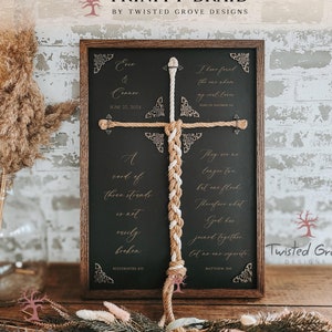 May include: A wooden framed sign with a black background and gold lettering. The sign features a trinity braid made of two ropes, one white and one brown. The sign includes the text "Ene & Conner" and the date "June 22, 2024". The sign also includes two bible verses: "I have found the one whom my soul love" and "They are no longer two, but one flesh. Therefore what God has joined together, let no one separate."