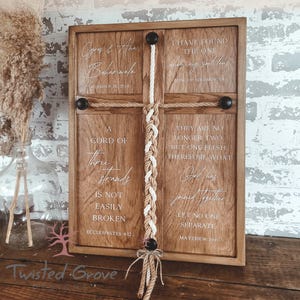 May include: Wooden framed wall art with a cross design. The cross is made of rope and features a braided center. The art includes text with names and dates, and Bible verses. The frame is brown.