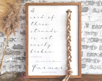 May include: A framed print with a white background and black text that reads "A cord of three strands is not easily broken" with the scripture reference "Ecclesiastes 4:12". Below the text is the couple's names "Jarrod & Katherine" and the word "Farmer" with the date "August 30, 2024". A braided rope hangs from the top of the frame.