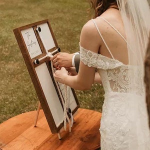 May include: A bride and groom take part in a wedding ceremony, tying a rope together on a wooden sign that reads "The Journey" with the date "07.10.20".
