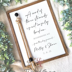 May include: A framed print with a white background and black calligraphy text that reads "A cord of three strands is not easily broken. Ecclesiastes 4:12 from this day forward Molly & Shane 2.11.23". A brown rope with white and tan strands is attached to the frame with black buttons.