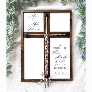 May include: A wooden frame with a white background and a brown rope tied in a braid. The rope is tied in a cross shape and has a black button at each end. The frame has the text "A cord of three strands is not easily broken" and "Ecclesiastes 4:12" printed on it. The frame also has the text "Adrian & Shyla April 24, 2022" printed on it.