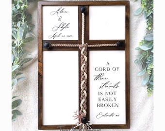 May include: A wooden frame with a white background and a brown rope tied in a braid. The rope is tied in a cross shape and has a black button at each end. The frame has the text "A cord of three strands is not easily broken" and "Ecclesiastes 4:12" printed on it. The frame also has the text "Adrian & Shyla April 24, 2022" printed on it.