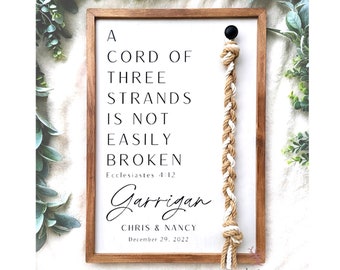 May include: A framed sign with a white background and a brown wooden frame. The sign features a quote from Ecclesiastes 4:12, "A cord of three strands is not easily broken." The sign also includes the text "Garrigan" with the names "Chris & Nancy" and the date "December 29, 2022". A knotted rope hangs from the top of the sign.