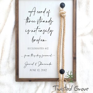 May include: A framed wooden sign with a white background and black text. The text reads "A cord of three strands is not easily broken."  The text "Ecclesiastes 4:12" is below the quote.  The text "from this day forward" is written in cursive below the verse.  The text "Jared & Sharanda" is written in cursive below the phrase.  The date "June 12, 2012" is written below the names.  A three-strand rope is attached to the sign with a knot at the top.  The rope is made of two natural colored strands and one white strand.  The sign has a black and white tree design in the bottom right corner.  The text "Twisted Grove Designs" is written below the tree design.