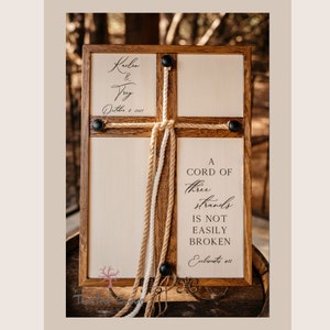 May include: Wooden framed sign with a cross design. The sign features the names "Kailen & Trey" and the date "October 1, 2021". The text "A cord of three strands is not easily broken" is also displayed. Decorative rope and black hardware complete the design.