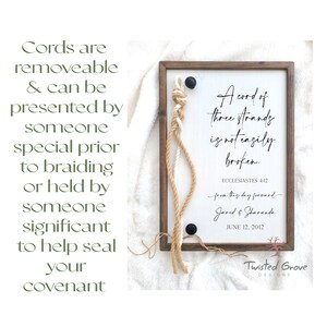 May include: A wooden sign with a white background and a brown frame. The sign features a quote from Ecclesiastes 4:12, "A cord of three strands is not easily broken." The quote is written in black cursive font. Below the quote, the text "from this day forward" is written in a smaller font. The names "Jared & Sharanda" are written below that, followed by the date "June 12, 2012". The sign is decorated with three cords, two brown and one white, that are tied together in a knot at the top. The cords are hanging down the sides of the sign. The sign is also decorated with a black metal button.