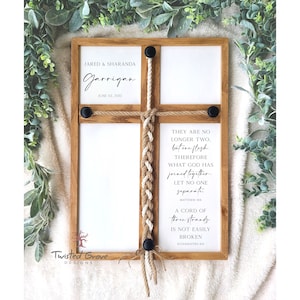 May include: A wooden framed wall art piece with a cross design. The frame holds four white panels, with text that reads "JARED & SHARANDA Jarrigan JUNE 02, 2012" and a bible verse. The cross is decorated with rope and black accents.