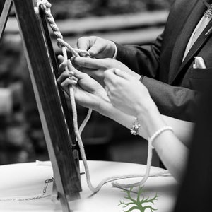 May include: A couple ties a knot in a white rope during a wedding ceremony. The rope is tied around a wooden frame. The couple is standing in front of a white table with a green tree logo and the text "Twisted Grove Designs".