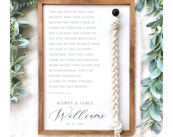 May include: A white wooden sign with a beige rope hanging from the top. The sign has a quote from Ecclesiastes 4:9-12 about the strength of unity. The sign is personalized with the names Audrey & James Williams and the date 08.21.2023. The sign is from Twisted Grove Designs.