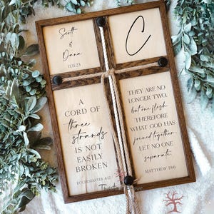 May include: A wooden sign with a rope braid in the center. The sign has the text "A cord of three strands is not easily broken" and "Ecclesiastes 4:12" on the left side. The right side has the text "They are no longer two, but one flesh. Therefore what God has joined together, let no one separate." and "Matthew 19:6". The sign also has the names "Scott & Dana" and the date "11.11.23" on the top left side.
