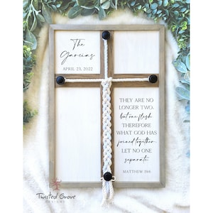 May include: A wooden sign with a white background and a rope cross in the center. The sign says "The Garcias" and "April 23, 2022" at the top. The rope cross is tied with a white and blue cord. The sign also includes the text "They are no longer two, but one flesh. Therefore what God has joined together, let no one separate. Matthew 19:6". The sign is framed with a light brown wood frame.