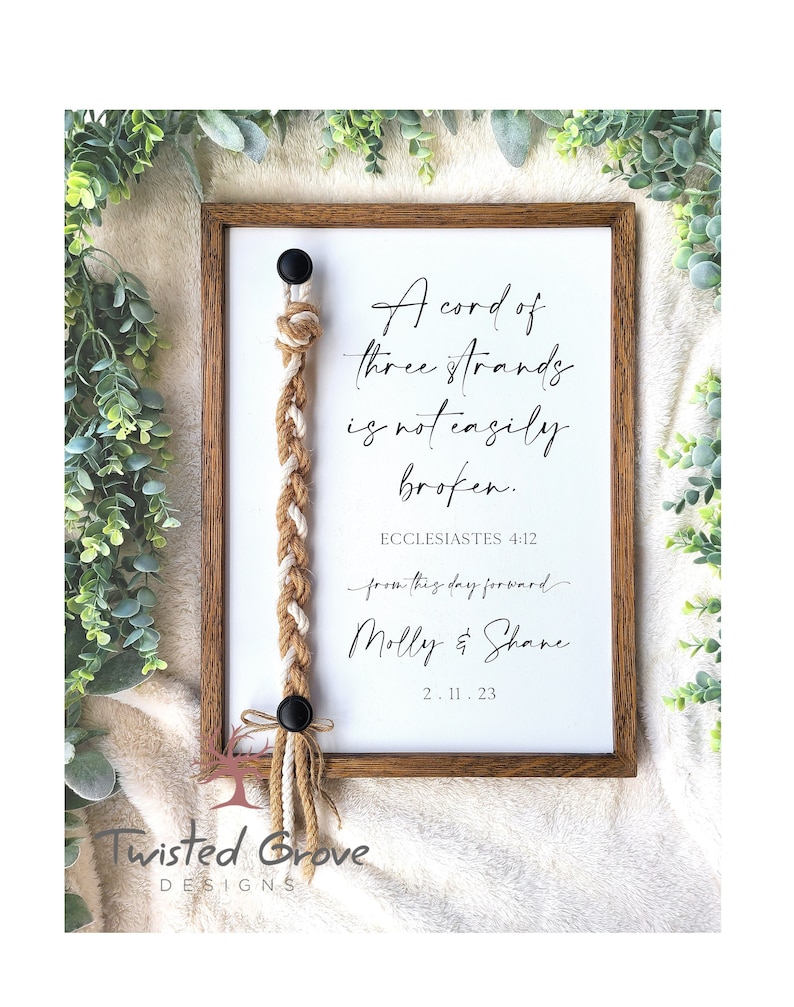 May include: A framed print with a white background and brown wood frame. The print features a braided rope with a quote from Ecclesiastes 4:12, "A cord of three strands is not easily broken." The print also includes the names "Molly & Shane" and the date "2.11.23".