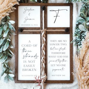 May include: A wooden sign with a white background and black text. The sign is divided into four sections. The top left section says "Joshua & Jamie" and "December 14, 2024". The top right section has a stylized letter "T". The bottom left section says "A cord of three strands is not easily broken" and "Ecclesiastes 4:12". The bottom right section says "They are no longer two, but one flesh. Therefore what God has joined together, let no one separate." and "Matthew 19:6". A white and brown rope is braided and tied to the sign.