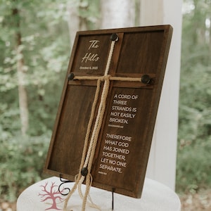 Peut inclure: Une pancarte en bois avec une corde nouée en forme de croix. La pancarte indique "The Hill's" et "October 6, 2023". La pancarte comprend également un verset biblique : "Une corde à trois brins ne se rompt pas facilement. Ecclésiaste 4:12. Par conséquent, ce que Dieu a uni, que l'homme ne le sépare pas. Marc 10:9". La pancarte est posée sur une table blanche avec un logo d'arbre qui dit "Twisted Grove Designs".