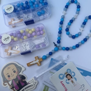 May include: Two clear plastic storage containers filled with blue, purple, and yellow beads, white cross charms, and wooden crucifixes. A completed rosary with blue and gray beads and a wooden crucifix, plus two illustrated cards featuring religious figures.
