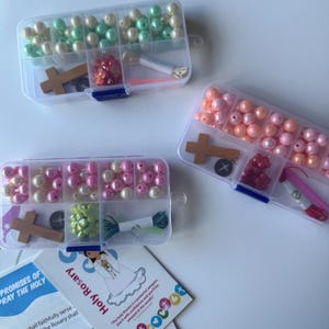 May include: Three clear plastic bead organizers filled with colorful beads, wooden crosses, and other rosary-making supplies. The organizers contain pink, peach, and green beads. A card with the text "Holy Rosary" is visible.