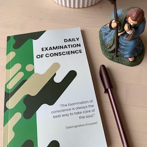 May include: A green and beige book with the title "Daily Examination of Conscience" and a quote from Saint Ignatius of Loyola. The book is on a wooden surface with a brown pen and a figurine of a person kneeling.
