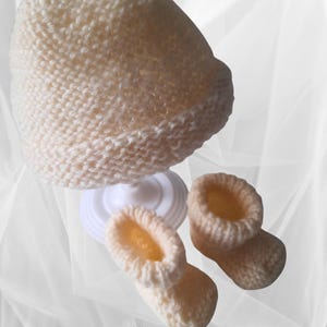 Peut inclure: Ensemble bonnet et chaussons pour bébé en tricot crème. Le bonnet est de forme arrondie avec un bord replié. Deux chaussons assortis sont également visibles. L'ensemble est en fil doux et texturé.