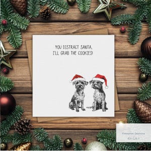 May include: A Christmas card featuring two terrier-like dogs in Santa hats. The card has the text "YOU DISTRACT SANTA, I'LL GRAB THE COOKIES!". The card is framed by festive decorations, including fir branches, baubles, and a star.